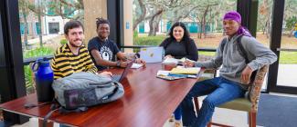 Four diverse students sitting at table with their books and laptops.